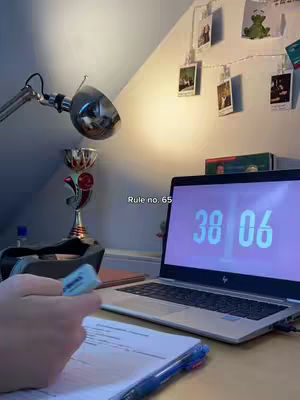 A person's hand holding a blue highlighter, over a notebook, and a laptop showing a clock. The laptop display shows the time of 38:06 in digital numbers. The room has a lamp on the corner, with photos attached to the wall.