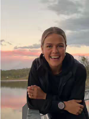 A smiling woman leans on a railing, against a backdrop of a sunset sky over water and trees. She is wearing a black jacket and a watch and appears happy and relaxed.