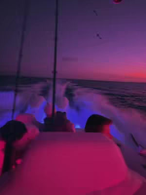 An eye-level shot from the back of a boat speeding across the water at sunset. The sky transitions from a deep purple near the horizon to a fiery red at the setting sun. The boat's interior is awash in shades of pink and blue from interior lights, with visible wakes and water spray.