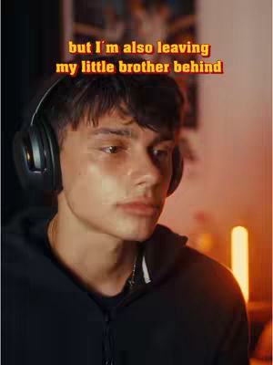 A young man with dark hair wearing a black hoodie and headphones looks downcast. The background is blurred, suggesting a room with warm lighting.