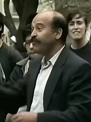 The image shows a man with a mustache in a dark suit jacket, light shirt, and open collar, speaking in an outdoor setting with other individuals in the background.