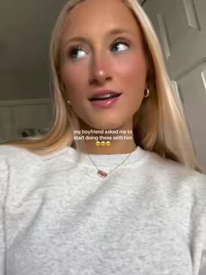 A young, blonde woman with light skin is the central focus, gazing slightly upward with a soft expression. She wears a light-colored, simple sweater and a delicate necklace. The background is slightly out of focus, suggesting an indoor setting with light-colored walls. A caption overlayed text at the bottom-center gives more info.