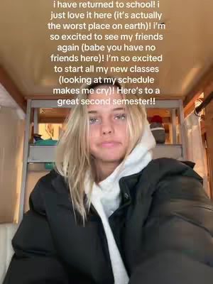 A young woman with long blonde hair is the central focus, captured from the chest up. She's dressed in a black puffer jacket over a white hooded sweatshirt. Behind her, a shelf holds various items, including a red cap. The background suggests an indoor setting, possibly a dorm room or bedroom, with the overall lighting appearing to be soft and natural.