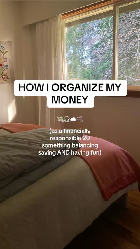 The image shows a cozy bedroom scene. A bed is made with a gray comforter and a red blanket. A window with sheer curtains and a view of trees is in the background. White text overlay in a white box reads, 'HOW I ORGANIZE MY MONEY' and a smaller text, (as a financially responsible 20 something balancing saving AND having fun).