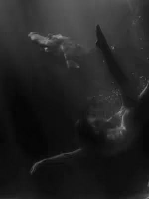 A grayscale image depicting a figure submerged underwater. The central focus is on the silhouetted form of a person, their body suggesting movement. The scene is predominantly dark, with subtle gradations of light highlighting the contours of the figure and suggesting the presence of water and bubbles. The composition is abstract, creating a sense of mystery and depth.