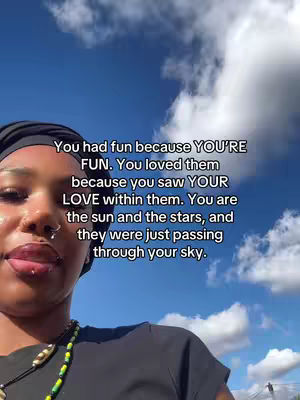 A selfie featuring a person with dark hair wearing a black top and a black head covering against a backdrop of a blue sky filled with white clouds. The person is positioned at the lower left of the frame. A text overlay is added.