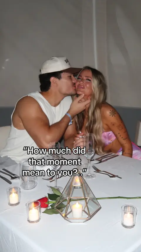 A couple kissing at a restaurant, with a table setting, candles, and a rose.