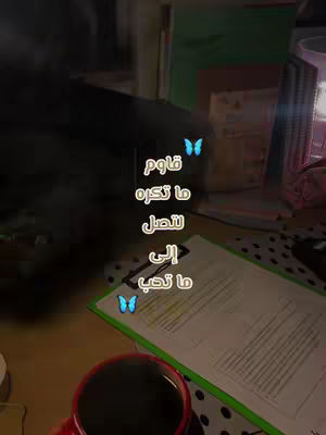 The cover image shows a desk with various items, including a coffee mug, a clipboard holding papers, and what appears to be a pen. The background is dimly lit and appears to be a room, with additional papers stacked on the side. The desk setup suggests a study or work environment. Overlayed on the image is text in Arabic, along with two butterfly icons.
