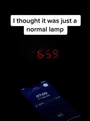 The cover image is a dark, nighttime scene. At the top, a white text box contains the phrase "I thought it was just a normal lamp". Beneath it, a digital clock is displaying the time as 6:59 in red digits. In the lower portion of the image, a smartphone screen is visible, displaying an alarm set for 7:00. The phone screen emits a blue glow, contrasting with the dark background.