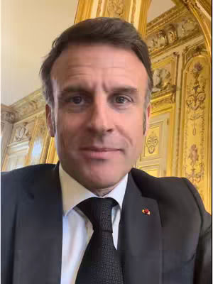 A selfie-style shot of a man in a suit and tie, appearing to be in a formal setting, likely an office. The background includes ornate gold-colored decorations suggesting a government building or similar environment.