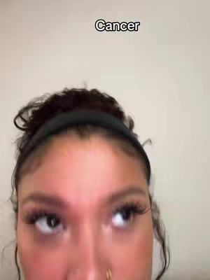 Close up shot of a woman's face, looking upward with a neutral expression. The word "Cancer" is printed in white text at the top of the image. The background is a plain, light-colored wall.