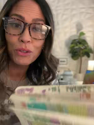 A woman with brown hair and glasses is the primary focus of the image, speaking directly into what appears to be a camera. The background is slightly blurred, suggesting an indoor setting, with hints of a decorative tree and wall. The woman holds a book or a similar item in her hands, partially obscuring the lower part of the frame. The color palette includes warm tones, enhancing the intimate feel of the video.