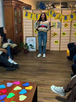 A woman stands in front of a screen divided into panels, with a banner reading 'BIENVENIDOS'. She is surrounded by people, a table of colorful hearts, and a sign. She is the focus of the image, looking at the camera and gesturing with her hands.
