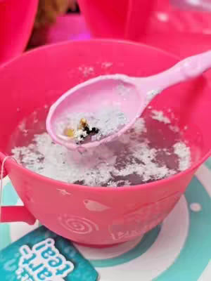A close-up shot of a pink teacup with a small spoon inside. The spoon holds a mixture of white and silver glitter with a few tiny, dark-colored objects. The cup is filled with a clear liquid, and there's a partially visible tea bag tag on the bottom left.