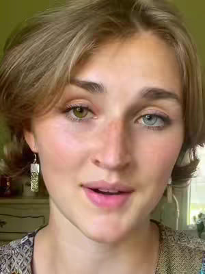 A medium shot of a young woman with an asymmetrical haircut and two different colored eyes. One eye is green and the other is blue. She has a natural makeup look, and is looking at the camera. The background is slightly blurred, but a light-colored dresser is visible.