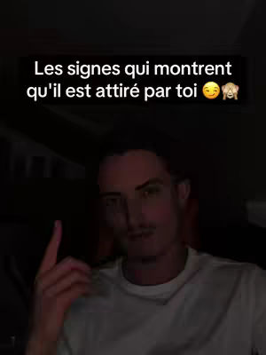 A man with short, dark hair and a mustache is the central figure, set against a dark backdrop. Above him, a text overlay reads, 'Les signes qui montrent qu'il est attiré par toi,' written in white font within a black rectangle, accompanied by a smiling face and hand-over-mouth emojis. The man is pointing his index finger upwards, suggesting a focus or emphasis on something.