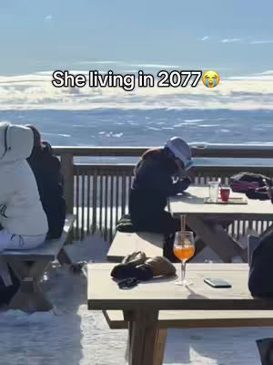 The image captures a person seated at a table with a view of a snow-covered landscape. The person is wearing dark clothing and a hat and is looking down. On the table, there is cocktail, a phone, and a pair of gloves.