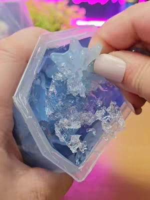 A close-up shot of hands manipulating a clear, hexagonal container filled with blue, gel-like substance and clear shards resembling ice. A hand is peeling away a piece of the gel-ice, revealing a star shape inside the container, while the background is blurred with hints of pink and green light.