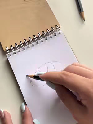 The image shows a close-up of a hand holding a pencil, drawing on a white notepad. The notepad is open on a wooden surface. The pencil is positioned to illustrate a drawing on the page. The drawing appears to be of an eye.