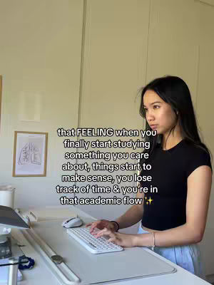 A young woman with long black hair sits at a desk with a laptop keyboard. On the screen is text in a white sans-serif font. The table is made of white. A framed picture is hanging on the wall.
