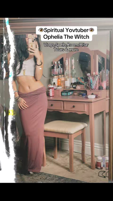 A woman stands in front of a mirror. She is wearing a white crop top and a long, flowing, mauve skirt. Her left hand is on her hip while the other holds a phone to take a selfie. Behind her stands a vanity with various beauty products and a mirror.