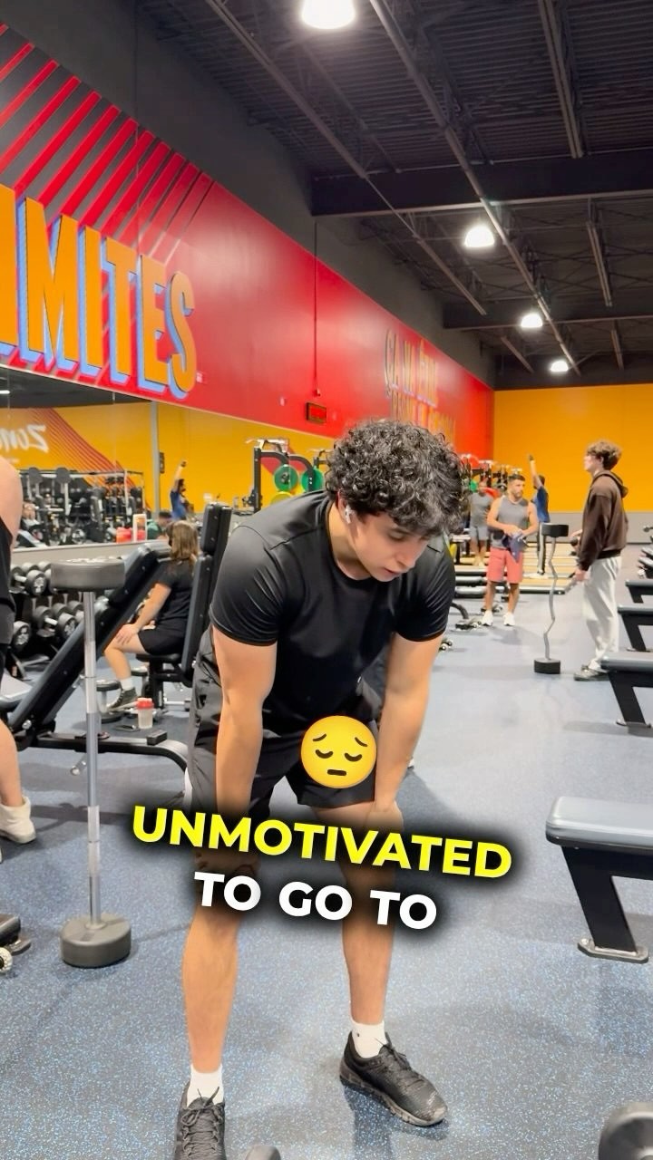 A young man in a black t-shirt and shorts is at a gym, hands on his knees, looking down with a tired expression. Text overlays read 'Unmotivated to go to...' in a yellow and white font. Gym equipment and other gym-goers are blurred in the background.