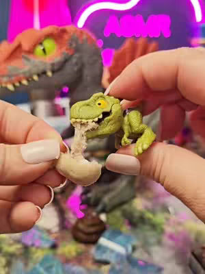 A pair of hands delicately hold a small Spinosaurus toy with a miniature rock-like substance emitting from its mouth. A second toy dinosaur is positioned in the background, set against a vibrant neon backdrop. The image has a shallow depth of field to create a sense of intimacy, and the close-up composition emphasizes the details of the toys and the hands involved.