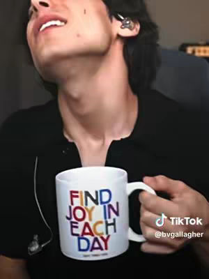 A person with dark hair and a black shirt is holding a white mug with the words "FIND JOY IN EACH DAY" written on it in various colors. The person has an earring and earpiece and is smiling. The background is slightly blurred.