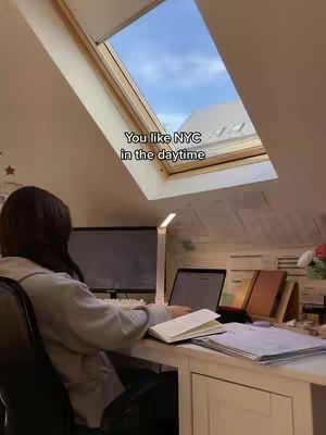 A woman sits at a desk near a window open to the sky. She is facing away from the camera, looking at a laptop and papers. The room is well-lit, with a modern desk and aesthetic decorations.