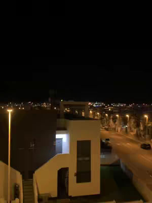 A nighttime cityscape, with a view from a high vantage point. The sky above is dark and clear. In the foreground, there is a building with a white wall and a staircase. Streetlights illuminate the scene, emphasizing a road and buildings in the distance.