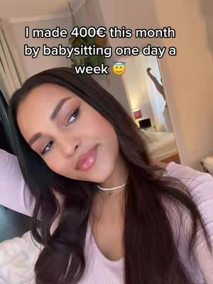 A woman with long dark hair is shown in a selfie-style shot, facing the camera with a slight smile. She wears a light pink top and a delicate necklace. Text overlays on the cover image indicate earnings from babysitting. The background reveals glimpses of an indoor setting, including a patterned curtain and possibly part of a bed in the blurred background.