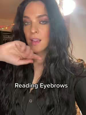 A woman with long dark hair is looking towards the camera. The text on the screen says 'Reading Eyebrows.' The background includes a room with a few lights.