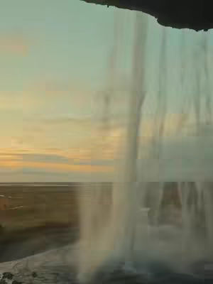 The image portrays a scenic view of a waterfall cascading down a cliff face, illuminated by the warm light of a sunset. The composition is divided roughly in half, with the top portion featuring the cliff and the sky painted in shades of orange and light blue. The bottom half is dominated by the waterfall's blurry flow and a field of green and brown below.