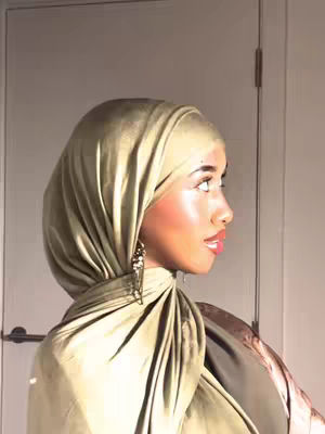 A woman wearing a green hijab is seen in profile against a light wall. The lighting highlights her face and the textures of the fabric.