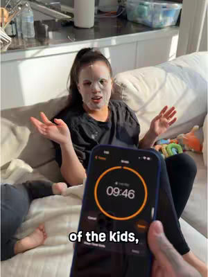 A woman wearing a face mask is sitting on a sofa with her arms open wide. She is looking towards the camera. In the foreground, a phone is held up showing a timer. The text "of the kids," is on the image. In the background, a child and part of their legs are lying on the couch.