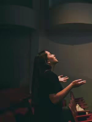 A woman in profile, looking up with her arms outstretched, in a dimly lit theater setting. The background includes rows of red seating and architectural curves.