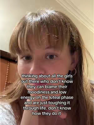 A selfie-style shot focuses on a woman's face. The background is soft, with neutral-toned walls and a glimpse of a dark wooden structure on the left, possibly a headboard or a piece of furniture. The woman has light-colored hair with a fringe. She is looking at the camera head-on with a slightly concerned expression. The overlaid text is white with a black outline and is in a plain font.