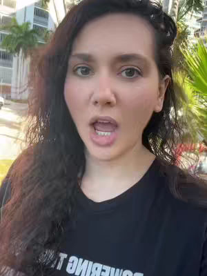 A medium shot of a woman with long dark hair, her mouth open in a surprised expression and the background showing a green, outdoor setting.