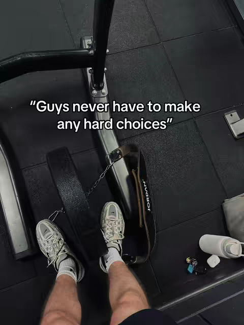 An overhead shot from someone's perspective in a gym. The gym is floored with black rubber tiles. The main focus is on the feet that are wearing sneakers, and a black weightlifting belt that's attached to the workout machine. Text overlaid on the image is in white letters. In the background are items such as a phone and a water bottle.