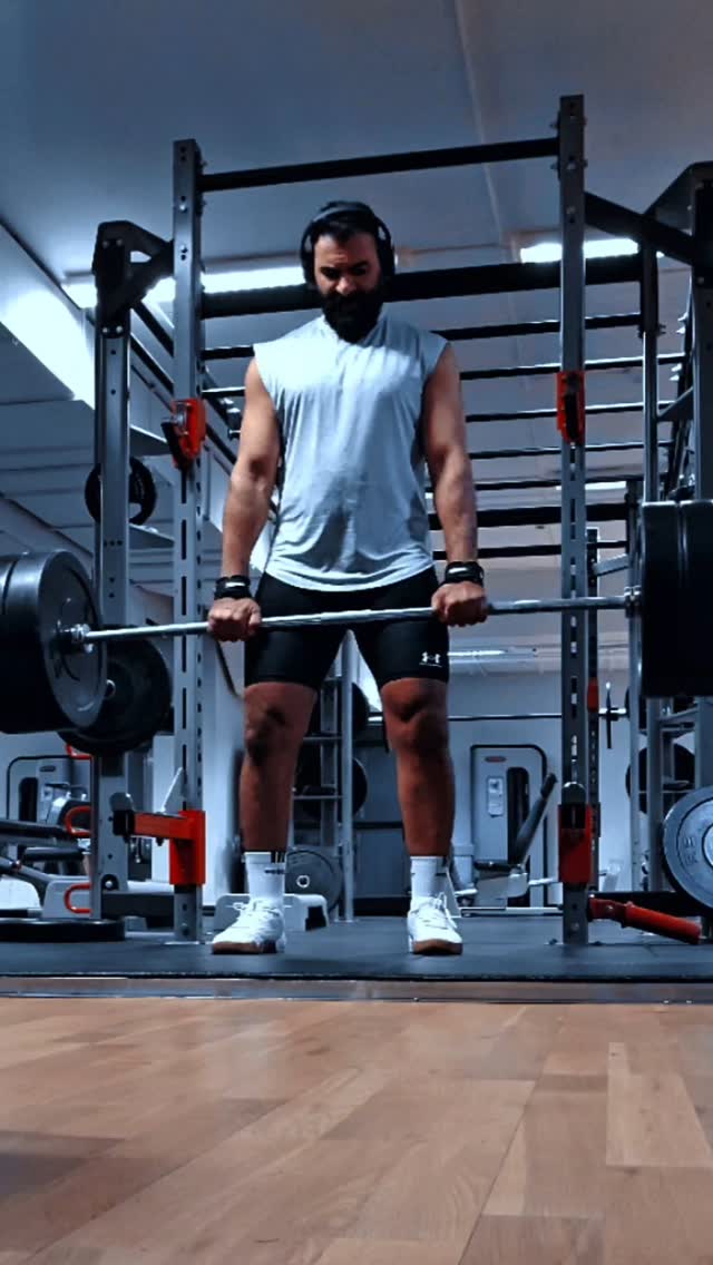 A man is lifting a barbell in a gym setting. He is wearing headphones, a sleeveless shirt, shorts, and wrist straps. Weights are loaded on the barbell, and the gym equipment is visible in the background.