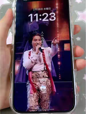 A person is holding a phone displaying a lock screen with a man on stage, holding a microphone, wearing a white shirt and patterned pants, and is pointing. The phone is in a clear case, and a hand is visible holding it. The background shows a colorful stage and the time.