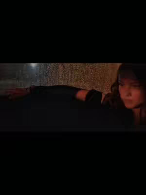 A person with long hair is reaching out towards a window or glass surface, possibly in the rain. The lighting is dim with a warm glow in the background and a dark foreground.