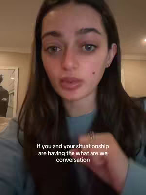 A young woman with long brown hair and fair skin is the focus of the image, positioned in the center. She's casually dressed in a blue long-sleeved shirt. Her expression is one of directness, looking straight at the camera, adding a conversational tone. Text overlayed at the bottom, featuring white font on a grey gradient background, reads: 'if you and your situation are having the what are we conversation'. The background is slightly blurred, revealing a room with wall art, suggesting an indoor setting.