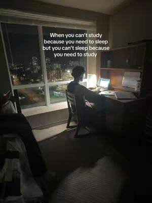 A dark, nighttime scene inside a room with a large window overlooking a city skyline. The room is dimly lit with a desk, a desk lamp, and a person sitting in a chair. The person is facing away from the camera, appearing to be studying. Text over the image reads "When you can't study because you need to sleep but you can't sleep because you need to study."