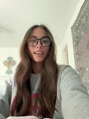A young woman with long brown hair and glasses is centered in the frame, speaking. She is wearing a gray sweatshirt. The background is a neutral-colored room with a white ceiling and a patterned tapestry hanging on the wall.