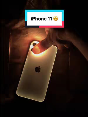 A dimly lit image featuring a hand holding an iPhone 11, with the back of the phone facing the camera. The phone glows with an internal light, possibly from a flashlight, causing the Apple logo to be visible. A text overlay at the top says iPhone 11.