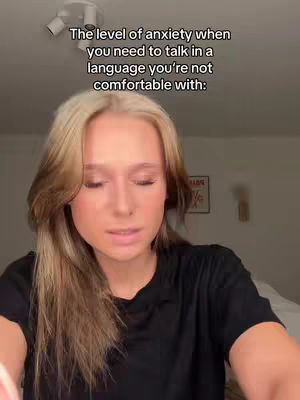 A young woman with long blonde hair is centered in the frame, looking down with a slightly concerned expression. Text overlayed at the top reads, "The level of anxiety when you need to talk in a language you're not comfortable with:" The background is a neutral, softly lit interior.