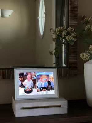 The image showcases a digital photo frame displayed on a white base, sitting on a dark surface. Behind the frame, a mirror reflects the room. The room appears to be well-lit, with a window and a vase of flowers adding to the ambiance. The frame displays two photos featuring groups of people.