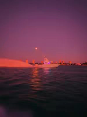 A boat is moving on the water at what looks like sunset, with a few buildings on the horizon. The colors are primarily deep pinks and purples.