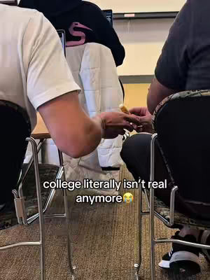 A low-angle shot from a seated position shows two people in what appears to be a college setting. The main focus is on the hands of the two individuals, one holding what seems to be a piece of food, possibly a snack. They are sitting on metal chairs. The background includes a table, jackets over chairs, with the overlay text: "college literally isn't real anymore".